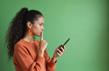 Obraz premium Thoughtful ethnic woman ponders message on mobile phone, finger near lips. Young African lady in casual orange jumper, large earrings considers online communication indoors against green background.