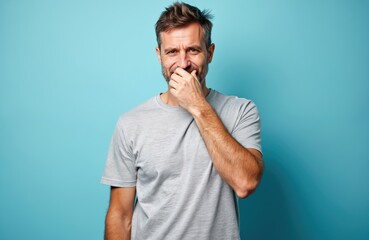 Middle-aged Caucasian man with beard covers mouth with hand, expressing surprise or secret. Adult male with blond hair stands against a vibrant blue studio background, looking worried or shy.