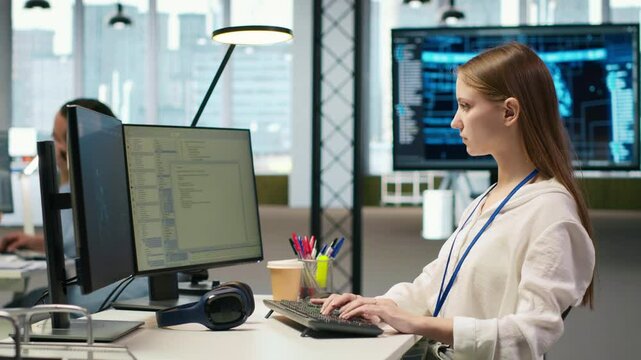 Female technician coding with AI language model assistance in startup office, using PC. Intern using computer, integrating artificial intelligence LLM assistant suggestions into code editor, camera A