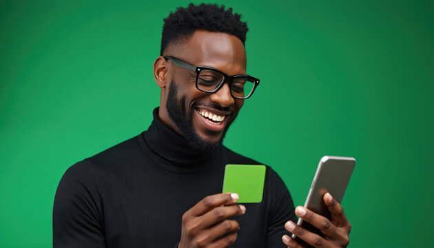 Happy Black man in glasses, black turtleneck uses green credit card for online shopping on smartphone. Smiling paying for purchases on mobile phone, enjoying e-commerce with digital banking.