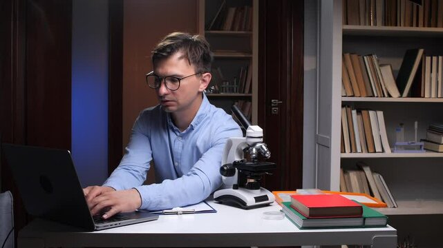 Scientist typing on laptop with microscope nearby in home office