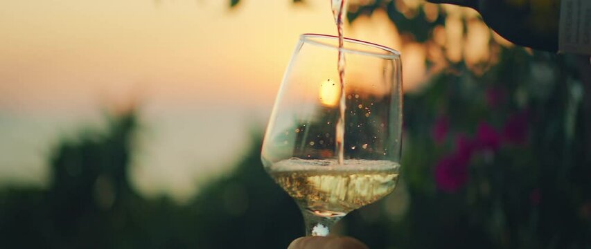 Close-up of white wine being poured into glass at sunset with sea and garden on the background