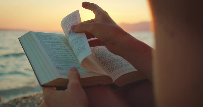 Woman reading Holy Bible book at sunset beach with pages turning and ocean view