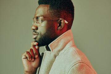 Thoughtful young man in stylish gray coat with glasses, posing against a soft green background,...
