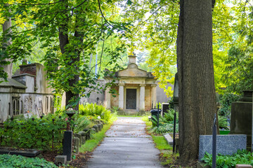 Tree-lined cemetery pathway leading to historic mausoleum building surrounded by verdant summer foliage creating serene memorial garden atmosphere