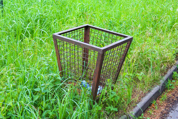 Metal mesh waste basket positioned among tall green grass in cemetery maintenance area creating...