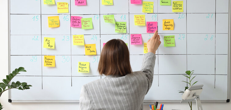 Young businesswoman sticking notes with plans on whiteboard in office, back view