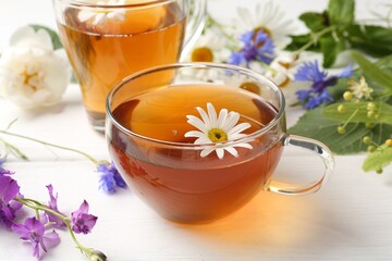 Aromatic herbal tea in glass cups, different flowers and green leaves on white wooden table, closeup