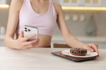 Woman with smartphone weighting donut on kitchen scale at white wooden table indoors, closeup