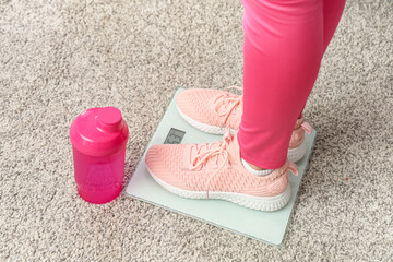 Beautiful young sporty woman with bottle of water standing on scales at home, closeup. Weight loss...