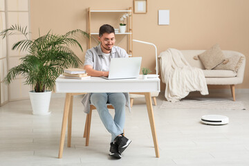 Young man using laptop at table with modern robot vacuum cleaner on floor in home office
