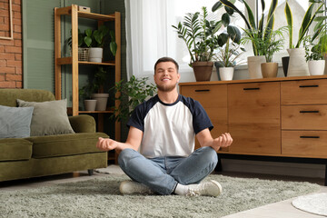 Young handsome man meditating on carpet in room with plants