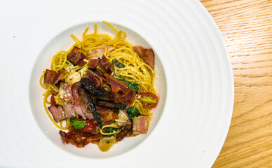 Flat lay of colorful pasta brunch with grilled bacon, herbs and veggies on white plate and wood table.