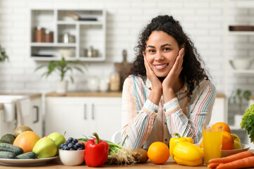 Young happy African-American woman with glass of juice, different vegetables and fruits in kitchen. Independence from Meat Day