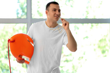 Happy male lifeguard with life board and whistle near window in pool