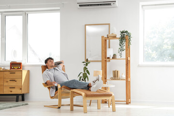 Young man with air conditioner remote control lying in armchair at home