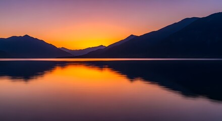 Obraz premium A vibrant sunset paints the sky orange and purple as it dips behind silhouetted mountains, reflected in the calm, still water of a lake.