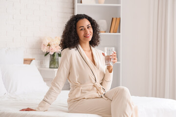 Young African-American woman with glass of water sitting in bedroom