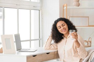 Young African-American woman with glass of water and laptop on table at home