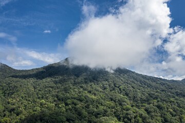 Lush green mountain peak shrouded in clouds against a blue sky in Jatiluwih, Bali, Indonesia. The cloud cover obscures the summit, creating a sense of mystery and natural beauty.
