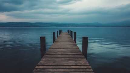 Obraz premium A wooden pier extends into a calm lake with a backdrop of distant mountains and a cloudy sky.
