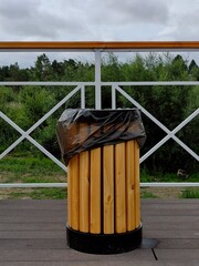 wooden trash bin with black liner sits on terrace near lush greenery. cloudy sky adds serene...