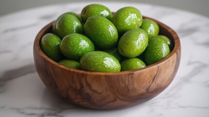 A wooden bowl overflowing with plump, green olives, sitting on a marble surface