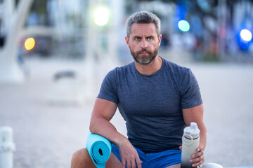 Sport fitness man resting on a bench outdoors. Mature fitness sport model sitting on a bench after a workout. Senior sports man taking a break after fitness. Fit guy resting after fitness.