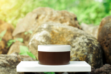 
A simple and elegant product mockup of a cosmetic jar with a blank label, placed on a white stand against a blurred natural background of rocks and green foliage. 