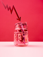 Fluctuating arrow shoots fall down at US dollars bill banknotes money cash inside glass jar on table, red tone background. Stock market crash, dollar weakens, business financial fall, money concept.