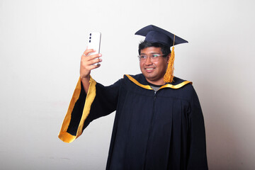 Young man dressed in cap and gown to celebrate his graduation uses his cell phone to take selfies,...