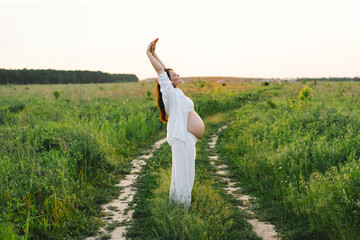 A pregnant woman stands in a green field. She gazes lovingly at her baby bump as the sun sets behind her, creating a serene atmosphere.