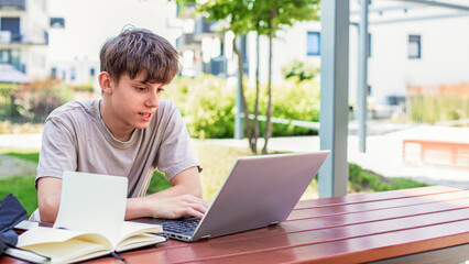Fototapeta premium Cute happy teenager with laptop studying online at table in yard, writing in notebook
