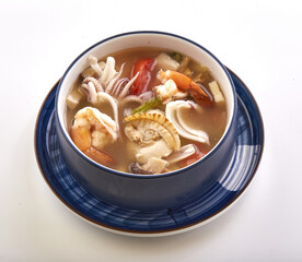 Bowl of spicy mixed seafood soup with shrimp, squid, crab, and vegetables in flavorful broth, served in blue ceramic bowl on matching plate. dish is vibrant and appetizing