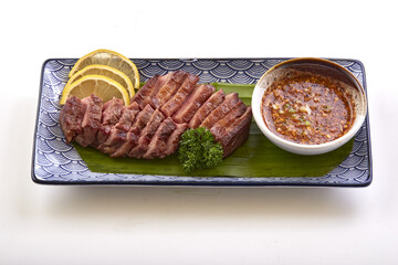Grilled beef tongue served on decorative plate with lemon slices, parsley garnish, and side of spicy dipping sauce, creating flavorful and appetizing presentation