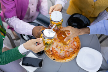 A group of friends cheers with drinks and pizza. The image is taken from above, showing the table...