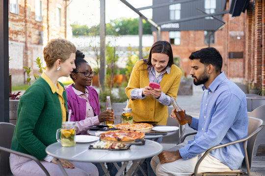 Diverse group of friends enjoying pizza and drinks together at an outdoor restaurant, smiling and using a phone. - Powered by Adobe