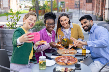 Friends enjoy pizza and take selfies at an outdoor restaurant, celebrating a happy occasion.