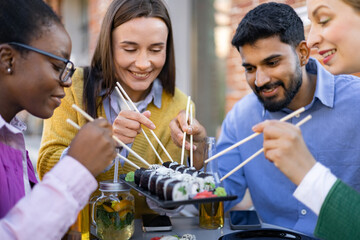 A diverse group of friends enjoy a meal together outdoors, savoring sushi and refreshing drinks.