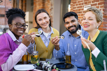 A diverse group of friends enjoys sushi together, smiling and laughing while eating outdoors on a sunny day.