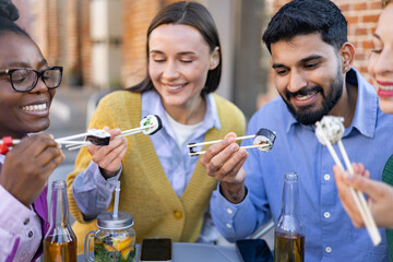 A diverse group of friends are enjoying sushi together outdoors, smiling and laughing while eating.