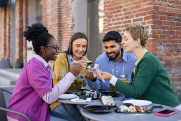 A diverse group of friends enjoys sushi outdoors, sharing food and laughter around a table.