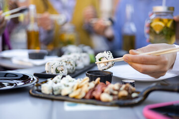Close-up of a hand using chopsticks to eat sushi from a table set with other food and drinks....