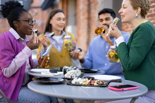 A diverse group enjoys sushi and drinks at an outdoor table, laughing and engaging in conversation. - Powered by Adobe