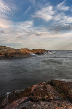 High-resolution landscape photo of a rugged coastline in Suomenlinna, Helsinki, Finland. The long exposure softens the movement of the sea against reddish coastal rocks