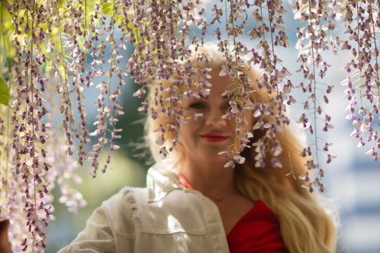 Wisteria Woman Portrait: Blonde girl smiles under purple flowers, garden outdoors in daytime for spring beauty