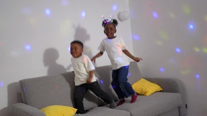 Two young children, a boy and a girl, are joyfully jumping on a gray sofa, illuminated by the colorful lights of a disco ball - Powered by Adobe