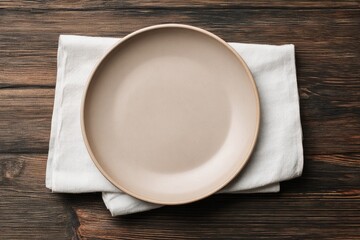 A taupe dinner plate rests on a folded white linen napkin atop a dark brown wooden surface