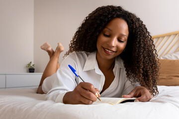 Pensive happy African American woman lying down on bed writing in personal journal in bedroom.