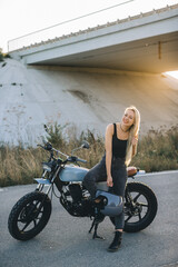 Smiling blonde biker posing with her motorcycle under a bridge